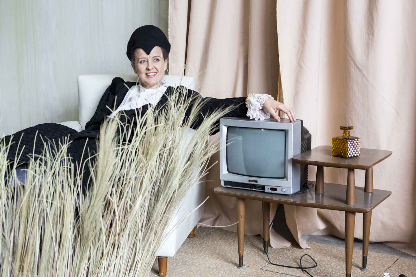 Taryn Fiebig on the set of Pinchgut's feature film, A Delicate Fire, sits in an armchair next to a retro TV, with dried grass in the foreground