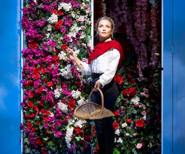 Soprano Alexander Oomens stands, holding a basket, in front of a wall of colourful flowers during a performance of Pinchgut Opera's Rinaldo
