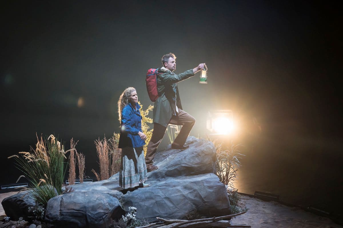 Sara Macliver as Eve wearing a blue jacket and Kyle Stegall as Adam wearing a green jacket and backpack, stand on a large rock set piece. Kyle holds a lantern as they both look off into the darkness.