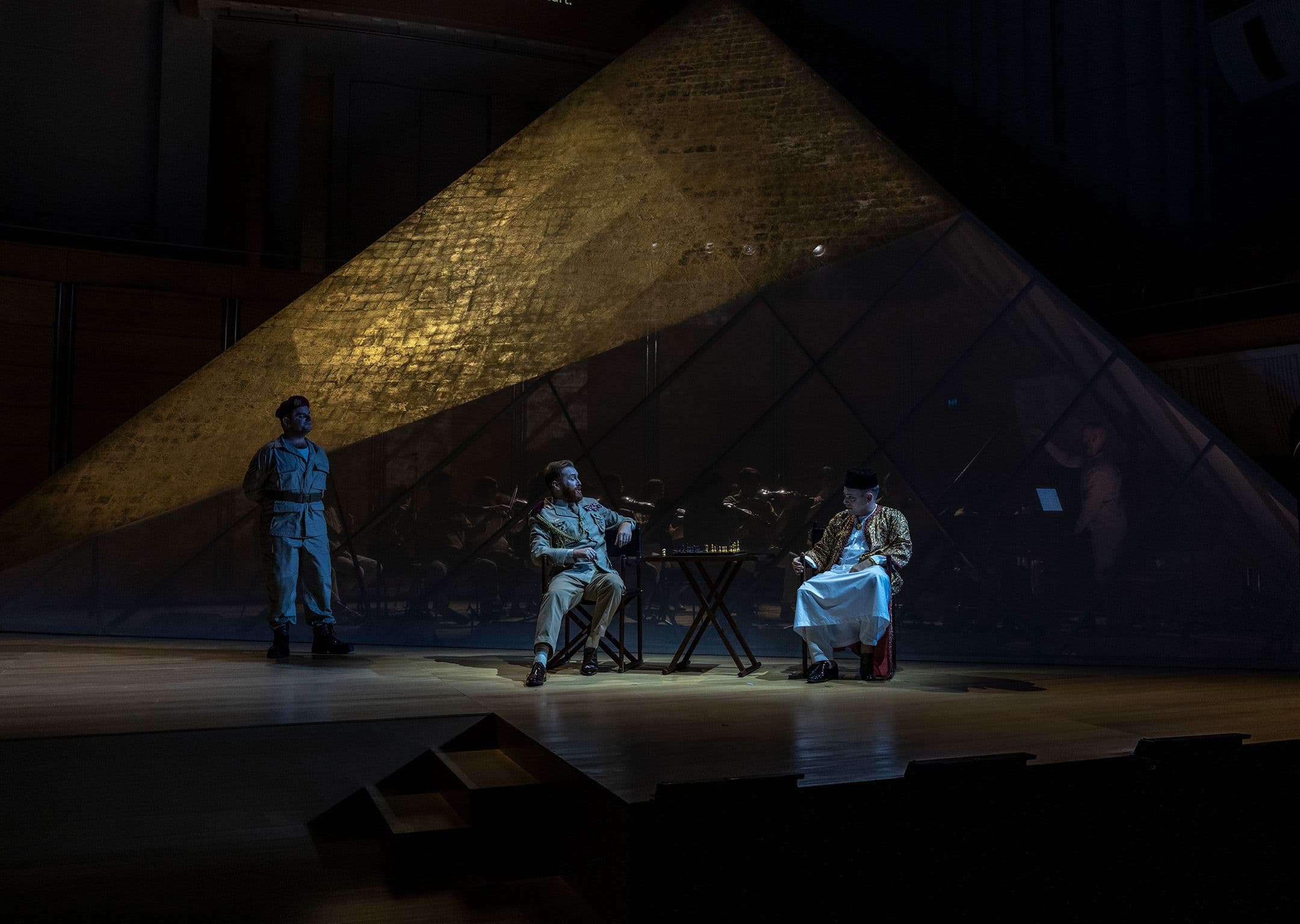 Countertenors Tim Mead and Hugh Cutting siting and playing chess in front of a large gauze pyramid on stage in Pinchgut Opera's production of Handel's Julius Caesar