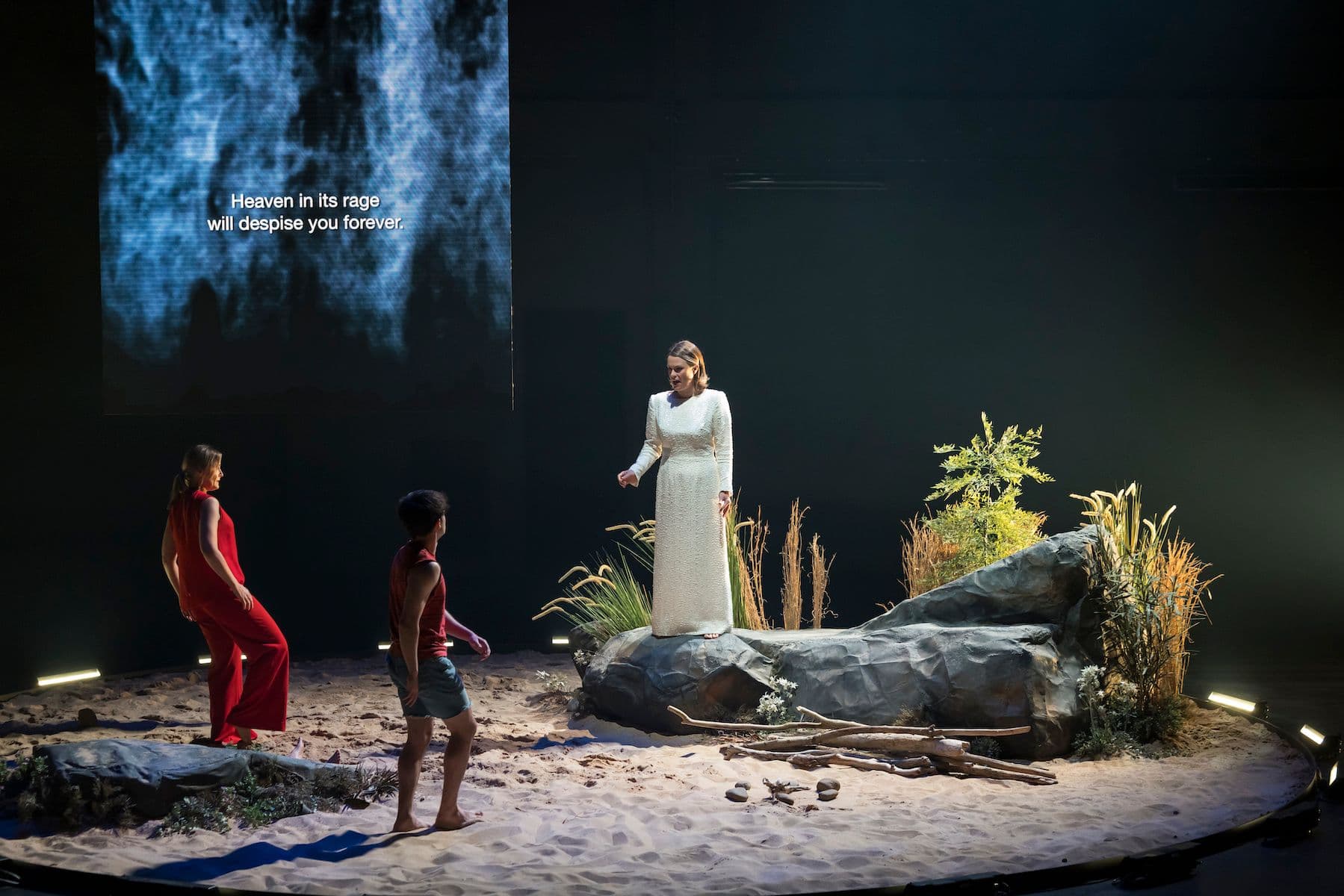 Stephanie Dillon as God, dressed in a full white gown-like dress, stands on a rock surrounded by sand, sings to Ashlyn Tymms and Ewan Herdman who both play Cain. They are dressed in red.